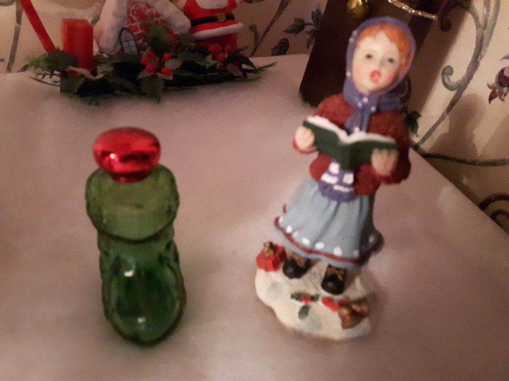 An image of a green glass boot perfume bottle with a red shiny top and a figurine of a little girl singing with a book in her hands.