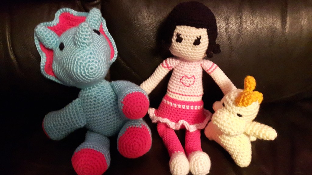 Three crocheted toys - a doll in a pink outfit and two dinosaurs. The large triceratops is blue with pink accents. The smaller dino is light yellow with dark yellow spikes down his back and tail